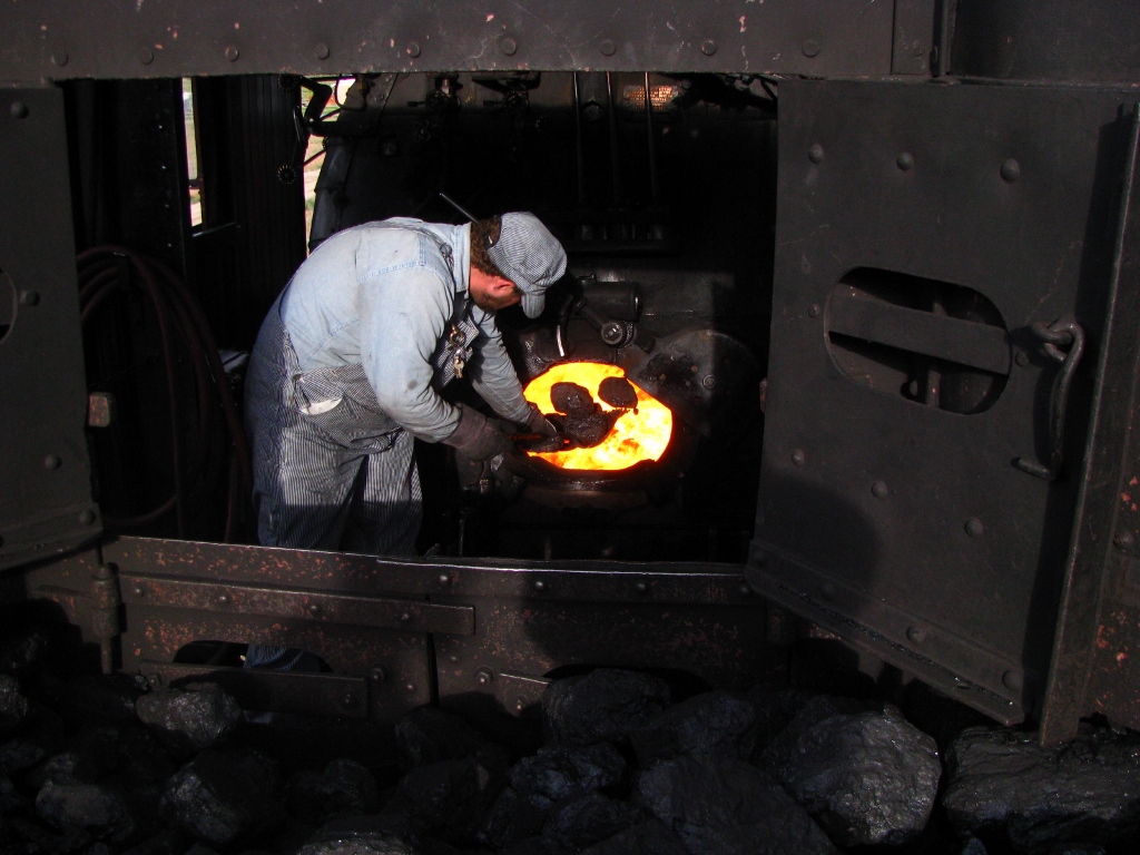 Railroad worker shovels coal lumps into a steam locomotive firebox.