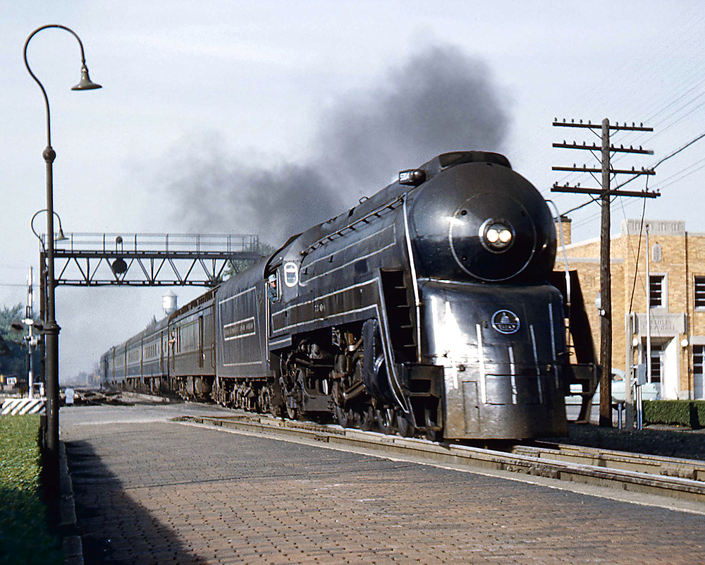 Color image of a bullet shaped steam locomotive at the head of a passenger train.