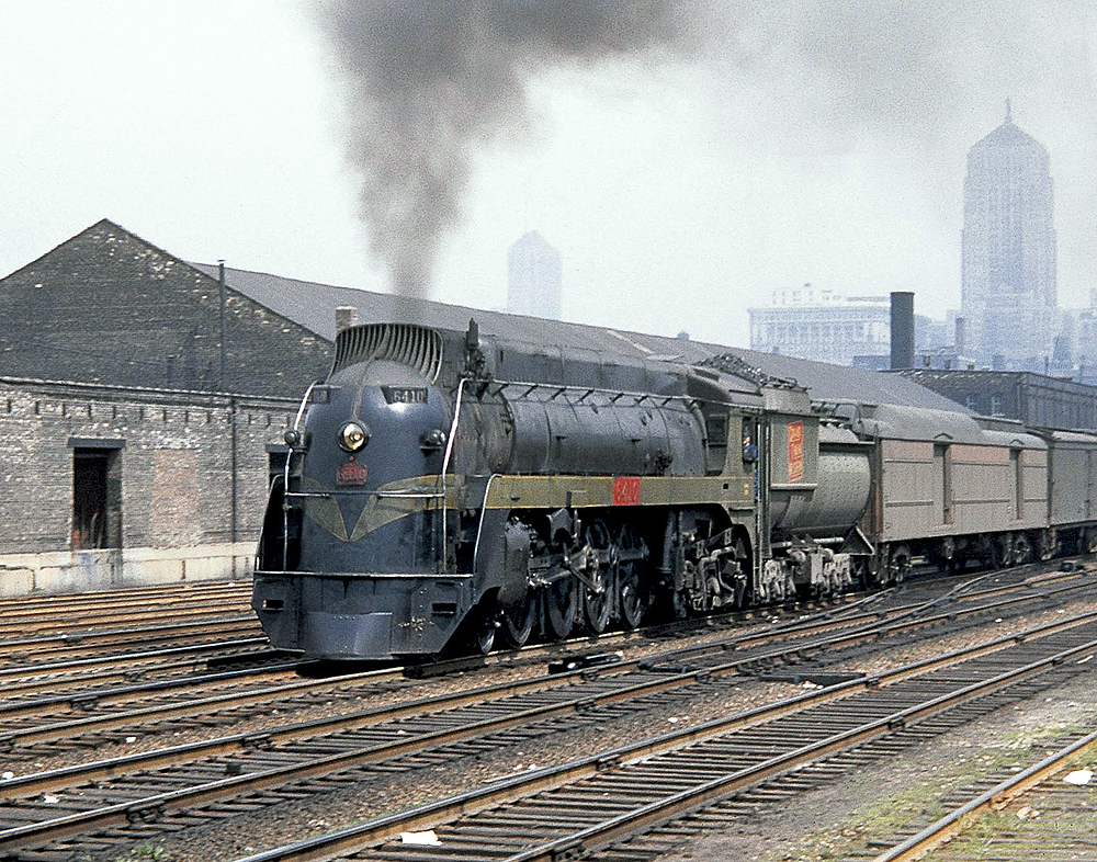 A steel gray steam locomotive at the head of a train in a rail yard.