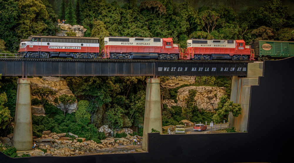 Three red-and-white Western Maryland diesels lead their train onto a girder bridge spanning a road and a creek