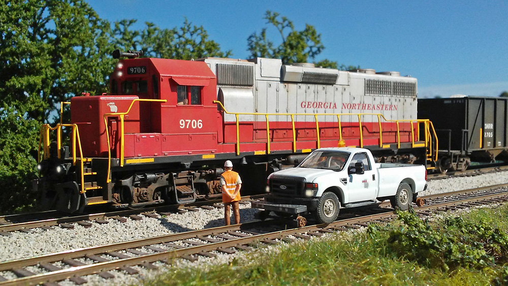 A railroad worker in an orange safety vest stands next to his Hi-Rail truck as he watches a red-and-silver diesel roll by on the other track