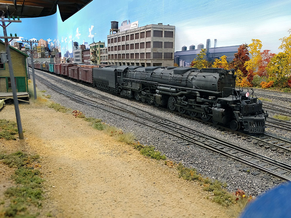 Union Pacific Big Boy moving left to right prepares to leave yard with mixed freight train, dirt rpad in foreground, industrial buildings in background