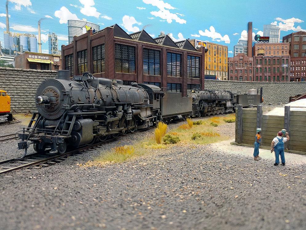 Santa Fe 2-10-2 faces left, yard crew stand in front of sand bin to right, brick car shop stands above locomotive in background