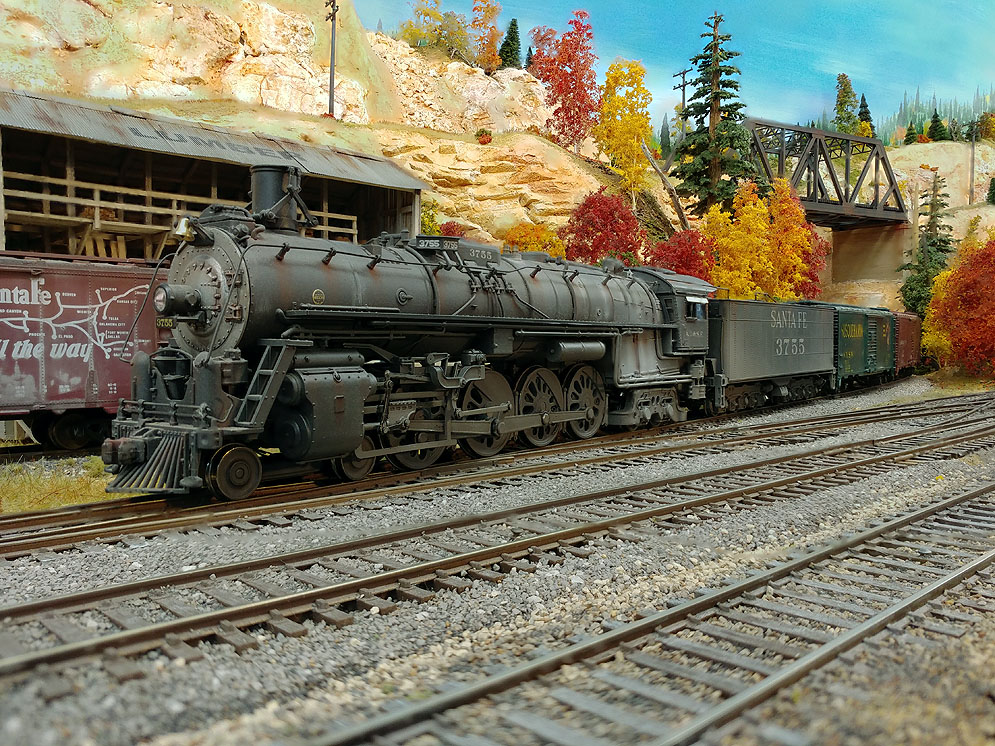 Santa Fe 4-8-4 facing left in front of partial Santa Fe boxcar with route map in front of wood-frame lumber shed, through truss bridge in upper right background