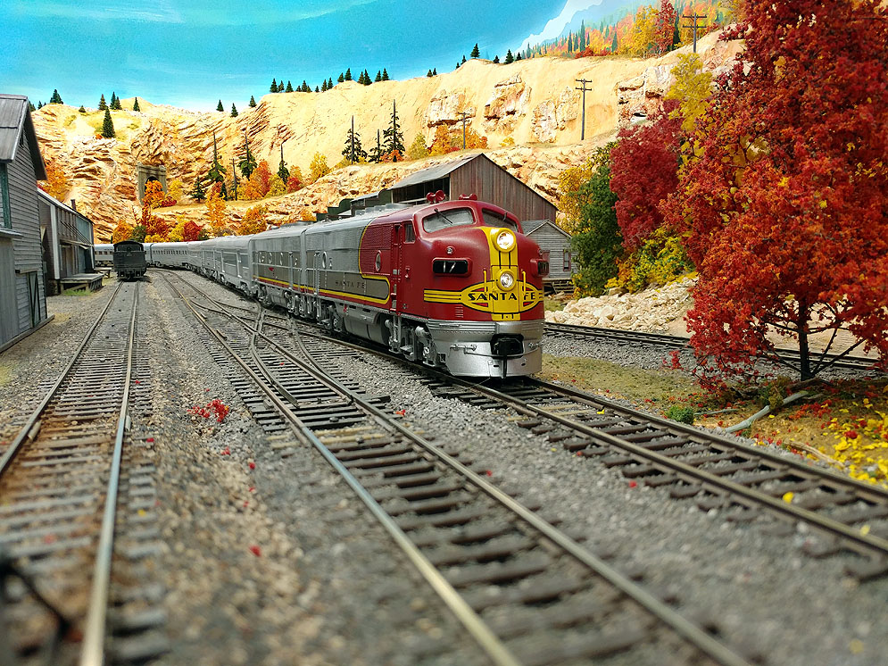Santa Fe F3 diesels in red and silver warbonnet scheme pull stainless stee passenger traind toward right along two-track main line through western mountain scene in fall