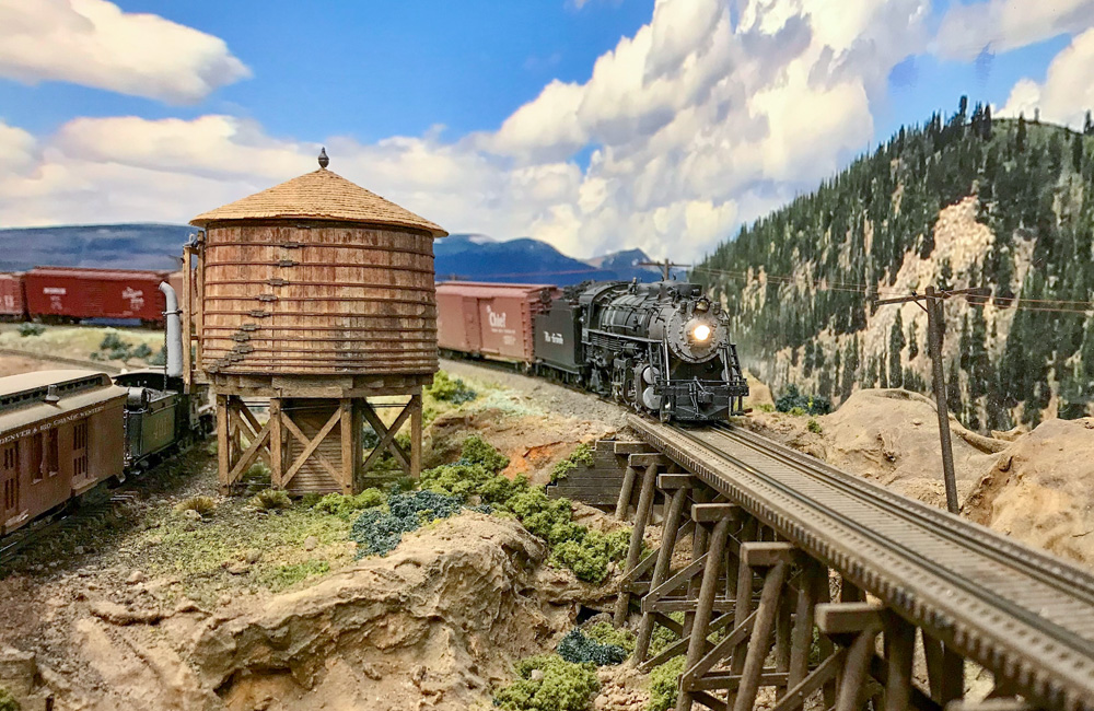 A steam locomotive approaches a trestle as another fills up at the water tank below
