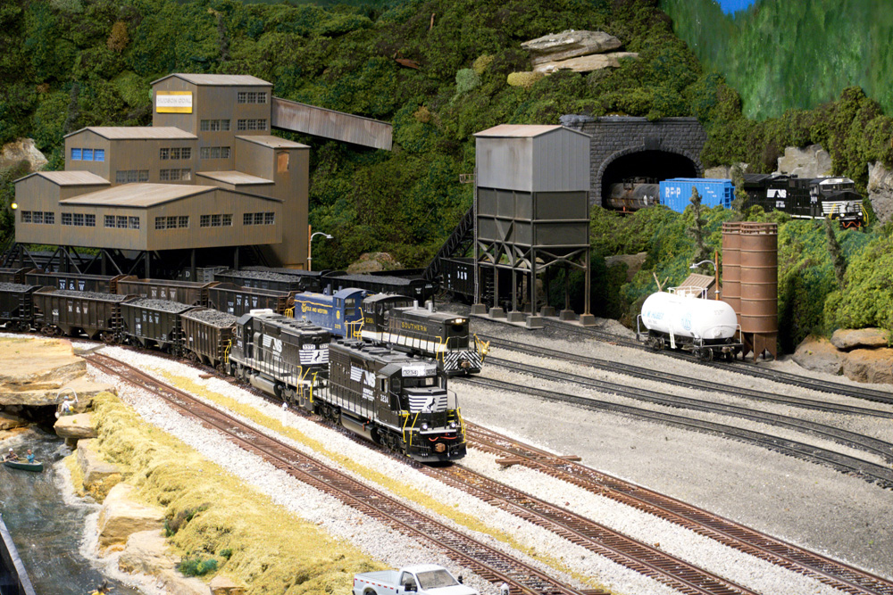 A pair of black diesels lead a loaded coal train while a diesel switcher works a mine in the background