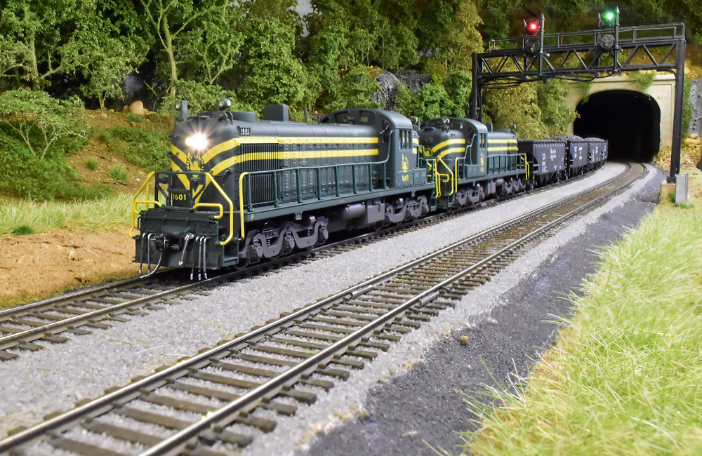 A pair of yellow-striped green diesels leads a coal train out of a tunnel under a forested hill