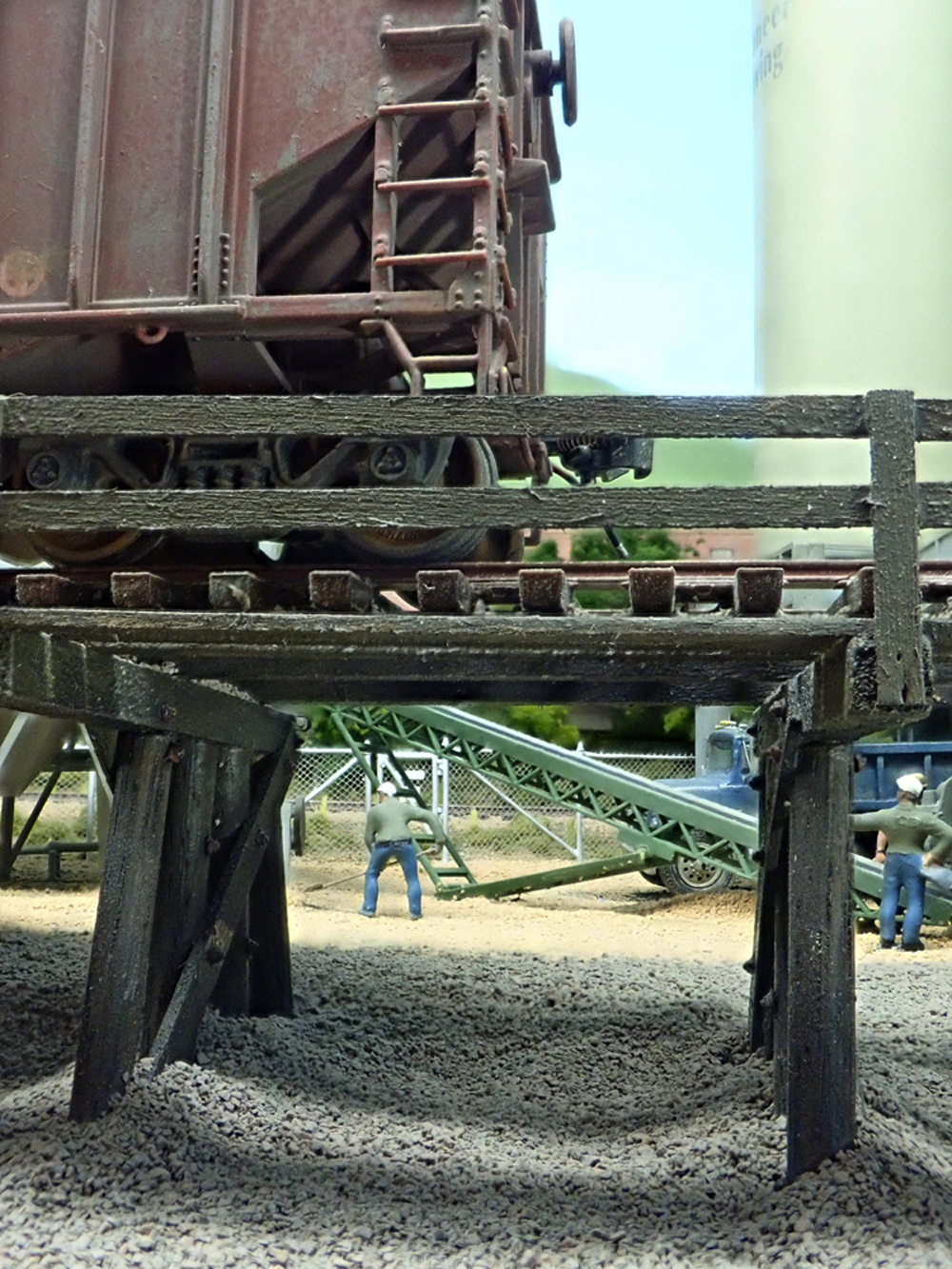 Two workers with shovels are framed by the legs of an unloading trestle bearing a gravel hopper