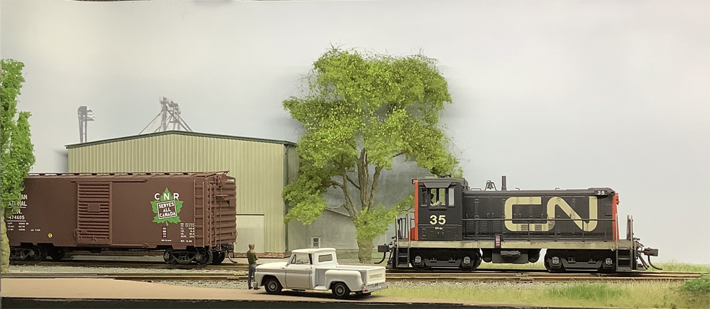 A black-and-red Canadian National switcher backs toward a brown CN boxcar in front of a corrugated metal building