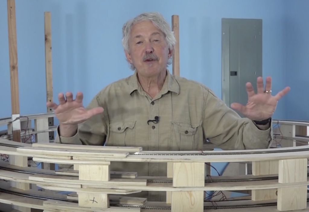 Older caucasian male gesturing with two hands while standing inside a model track helix.