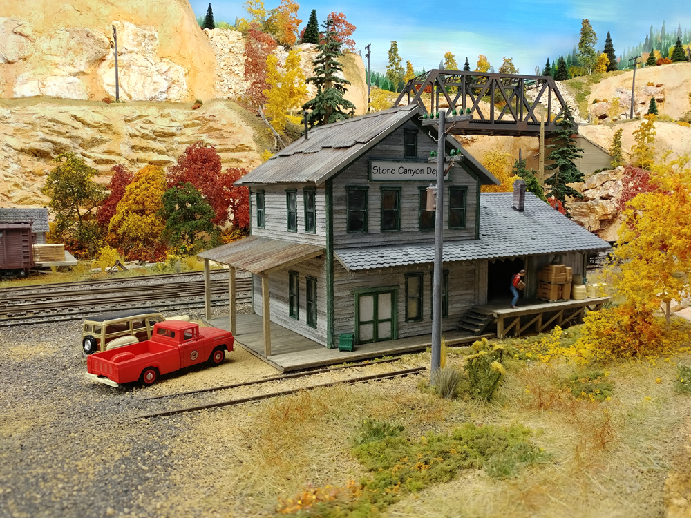 Gray wooden two-story structure with red Ford pickup and tan 1940s Ford Woody station wagon parked in gravel lot to its left