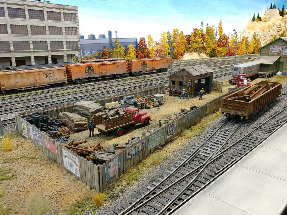 Wood fence encloses junkyard with red stakebed truck and corrugated steel office, rusty gondola spotted at siding