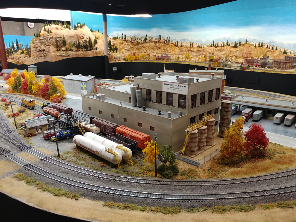 Gray two-story masonry structure with two-track siding holding boxcars and tank cars, two white propane tanks parallel siding, curved main line passes in front of scene