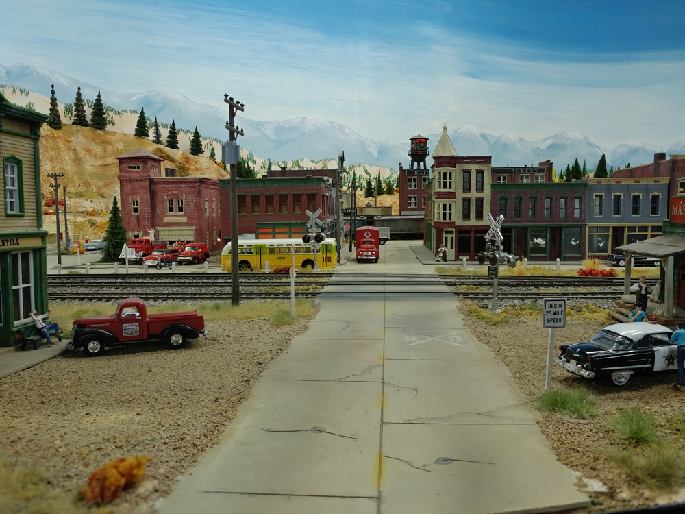 Cracked concrete street crosses two-track main line before heading into downtown area with storefronts to the right and brick firehouse to the left, black and white police car in right foreground