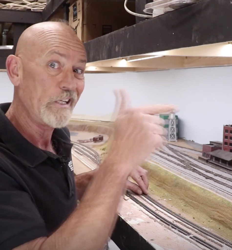 Older caucasian mail with shaved head gesturing and making a face while pointing at his model railroad layout.