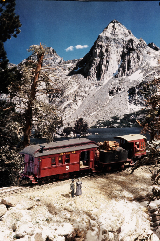 Two formally dressed figures stand beside a steam-powered one-car excursion train in a mountainous setting