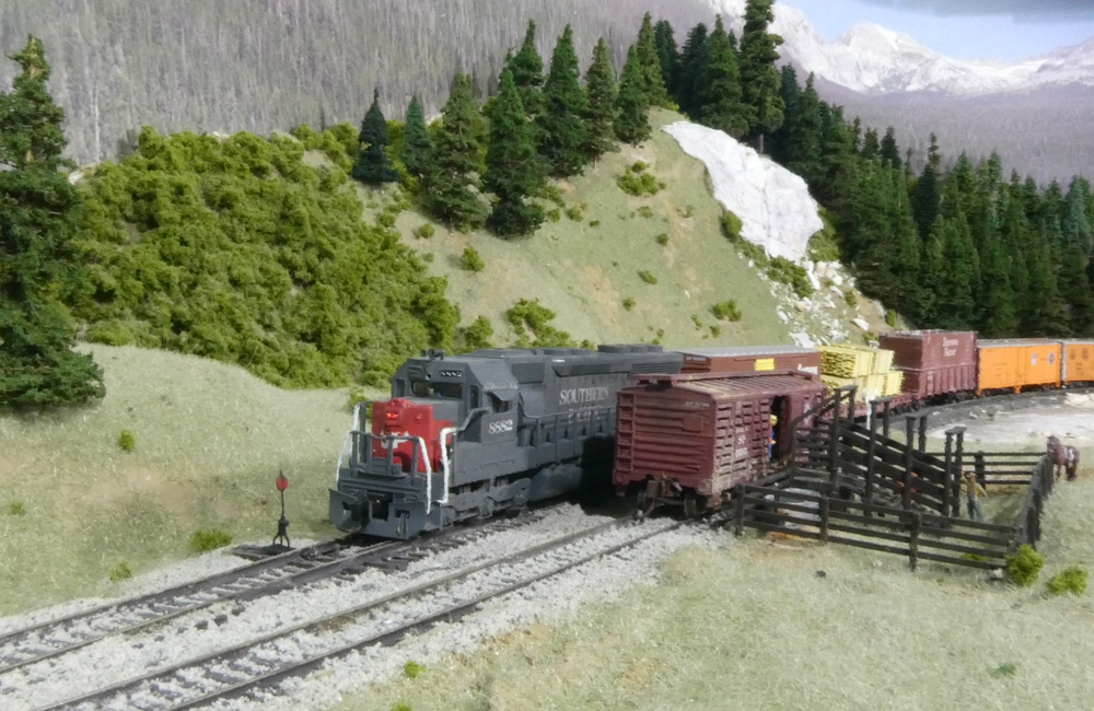 A gray-and-scarlet diesel with a manifest train passes by a stock pen in the mountains