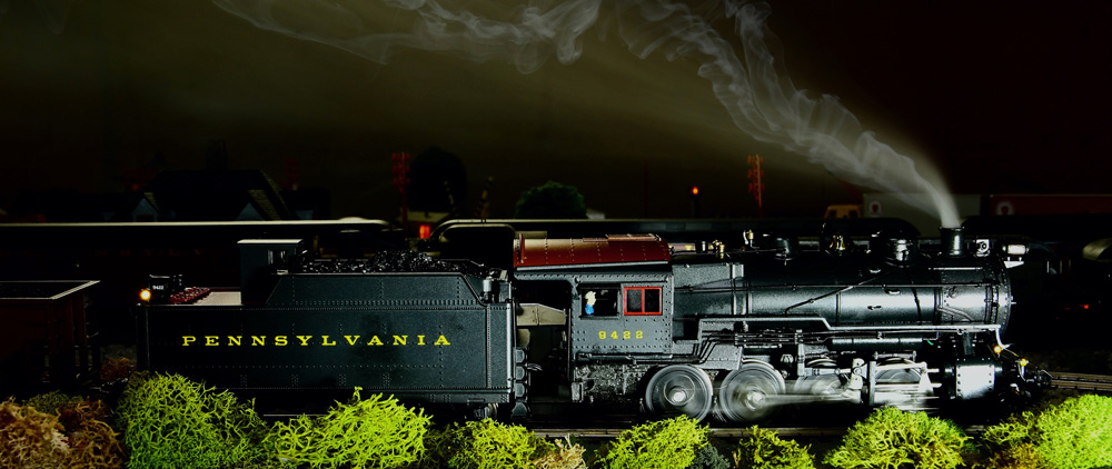 A steam engine, wheels blurred by motion, is illuminated by camera flash as it rushes by at night