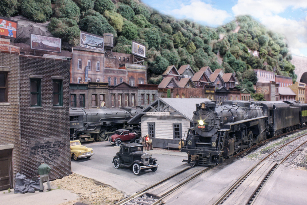 A weathered steam engine leads a train past a white clapboard station in an Appalachian town