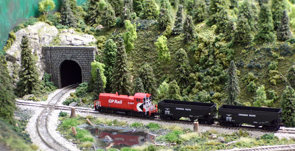 A red-and-black Canadian Pacific diesel switcher pulls a pair of empty hoppers in a wooded setting