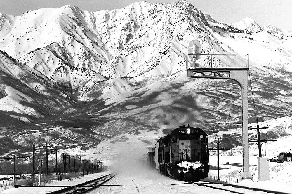 Freight train kicking up snow under a signal gantry