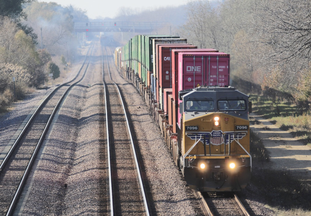 Yellow locomotive pulling stacked container cars on undulating straight track. 