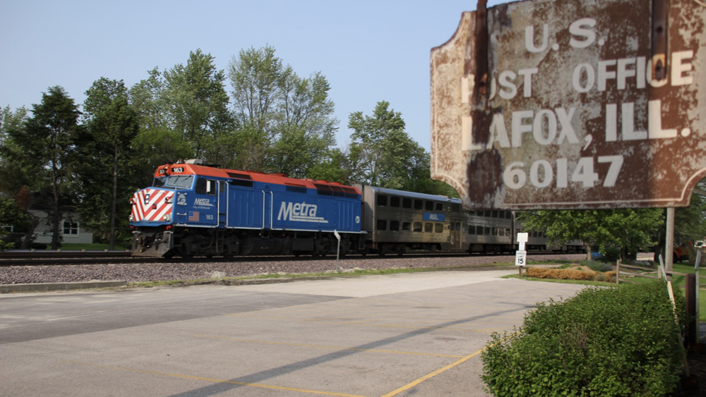 Commuter train with blue locomotive passes in background with post office sign in foreground.