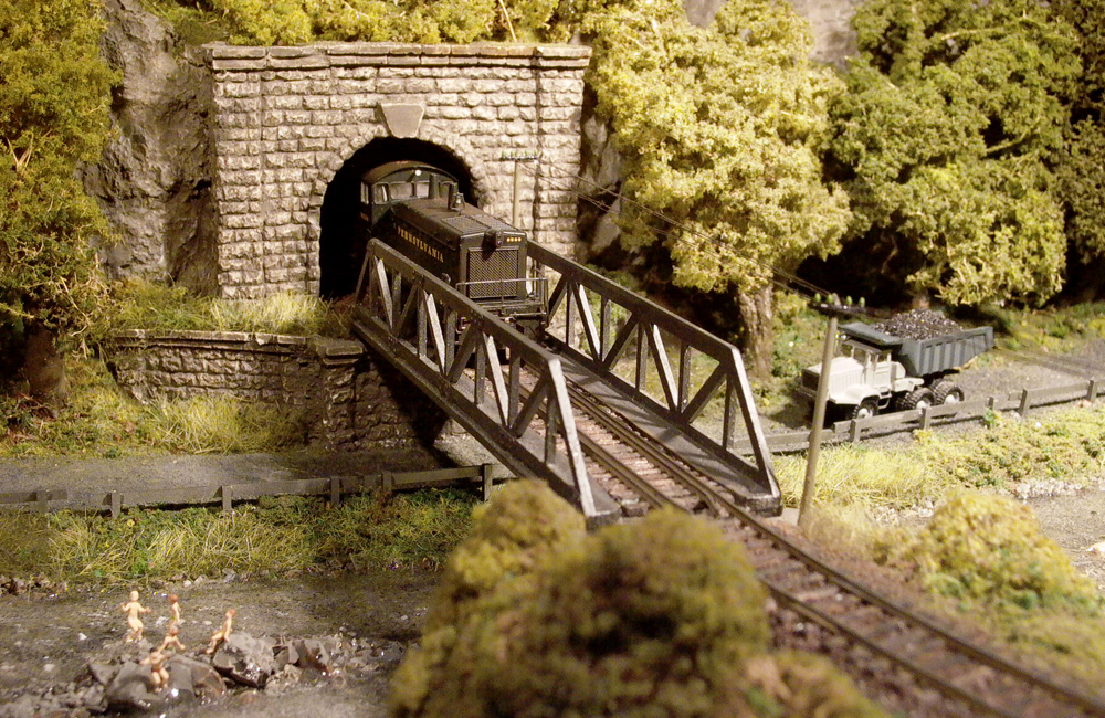 A Pennsylvania Railroad diesel switcher emerges from a tunnel onto a pony truss bridge above a road and creek
