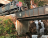 A heavily weathered Southern Pacific diesel locomotive emerges from a tunnel onto a plate girder bridge over a waterfalling creek