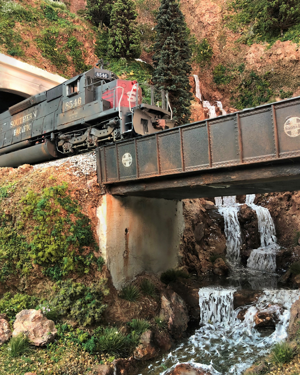 A heavily weathered Southern Pacific diesel locomotive emerges from a tunnel onto a plate girder bridge over a waterfalling creek