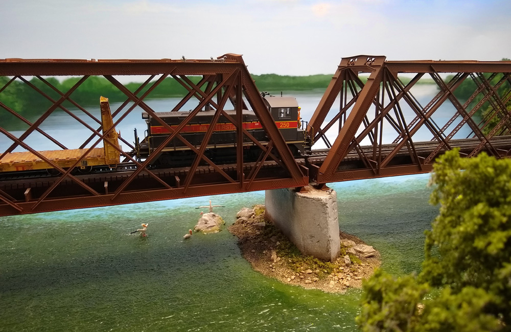 A red-and-black diesel switcher running cab-end-first pulls a yellow bulkhead flatcar over a bridge
