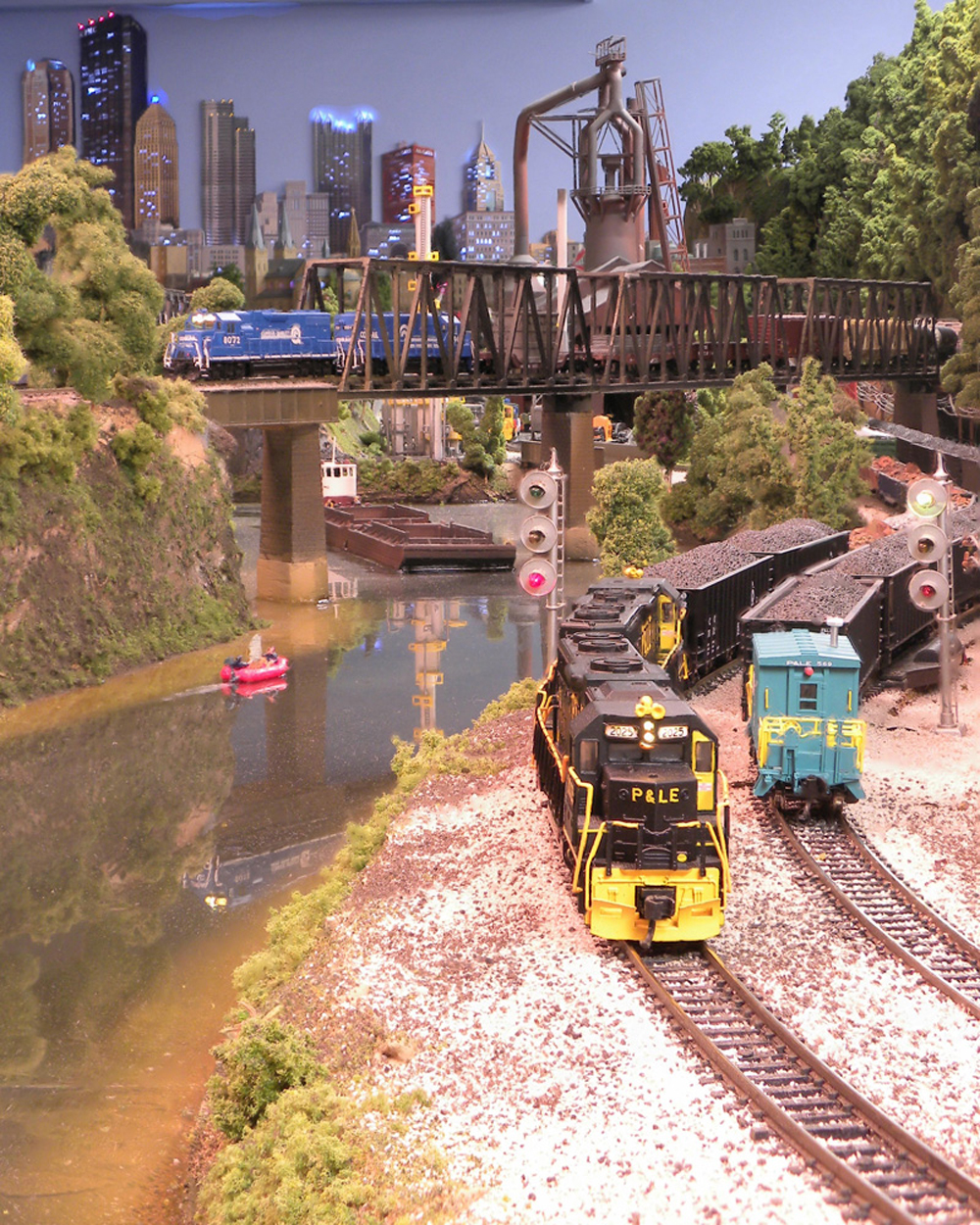 A pair of coal trains wind under a bridge and along a river toward the camera with a city scene in the background