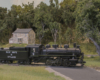 An 0-6-0 steam locomotive pulls a modern boxcar across a road crossing in front of a fieldstone farmhouse