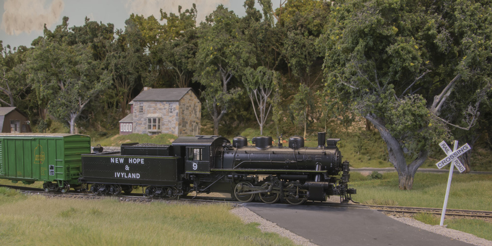 An 0-6-0 steam locomotive pulls a modern boxcar across a road crossing in front of a fieldstone farmhouse