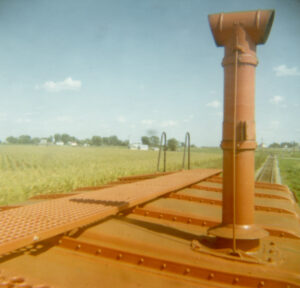 Orange-red smokestack, roof walk, and a ladder end appear in the foreground against a backdrop of row crops and blue sky filled with distant fluffy clouds.