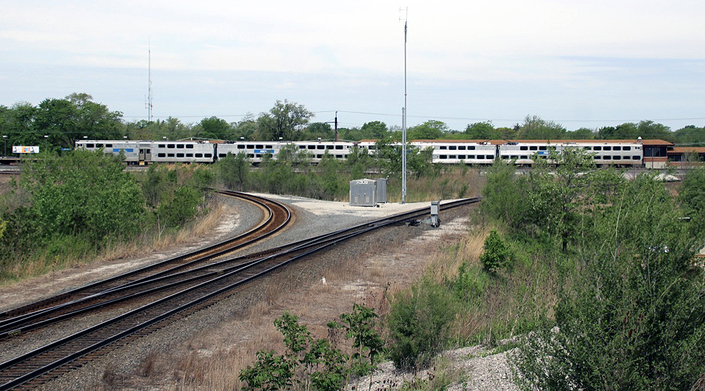 Four-car stainless steel electrified commuter train, partially obscured by brush, stops at station.