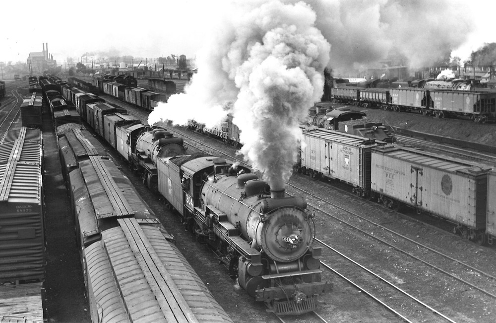 Black-and-white overhead view of steam locomotive with freight train in freight yard