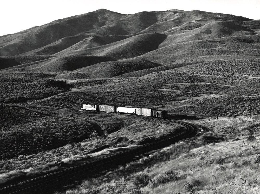 Black-and-white view of road-switcher diesel locomotive with short freight train amid barren hills