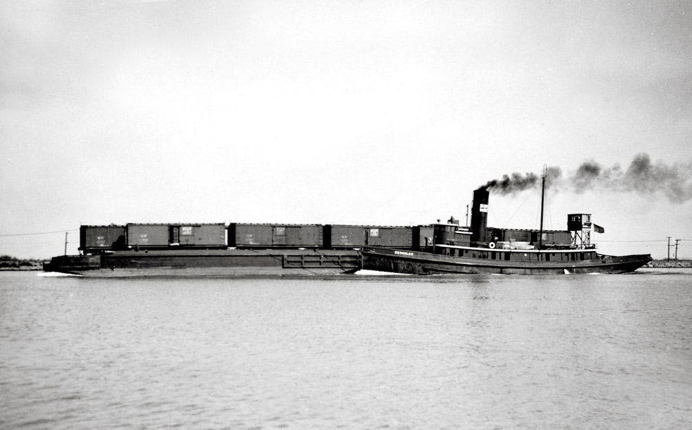 Black-and-white broadside view of tugboat with barge carrying freight cars