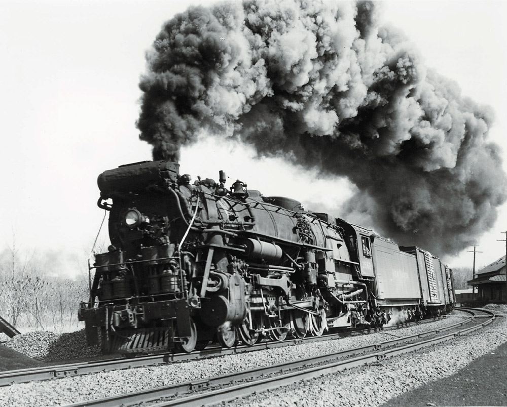 Low-angle black-and-white photo of 4-8-2 steam locomotive making smoke with freight train