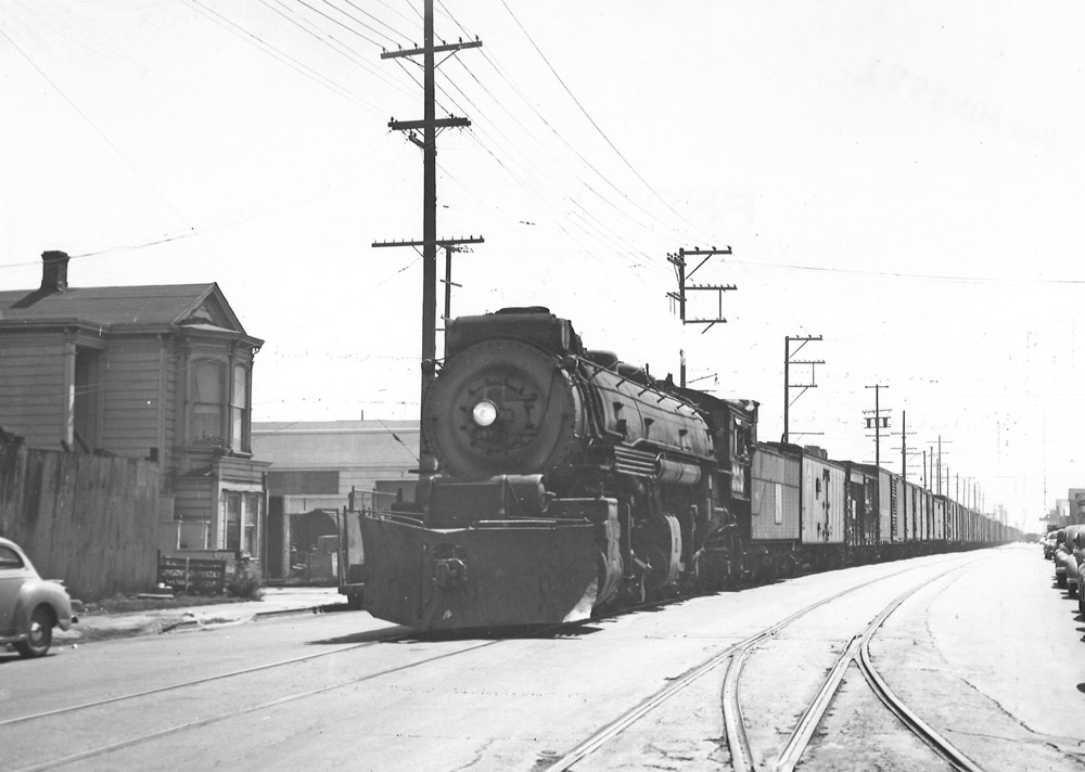 Black-and-white view of steam locomotive with freight train on track in a city street