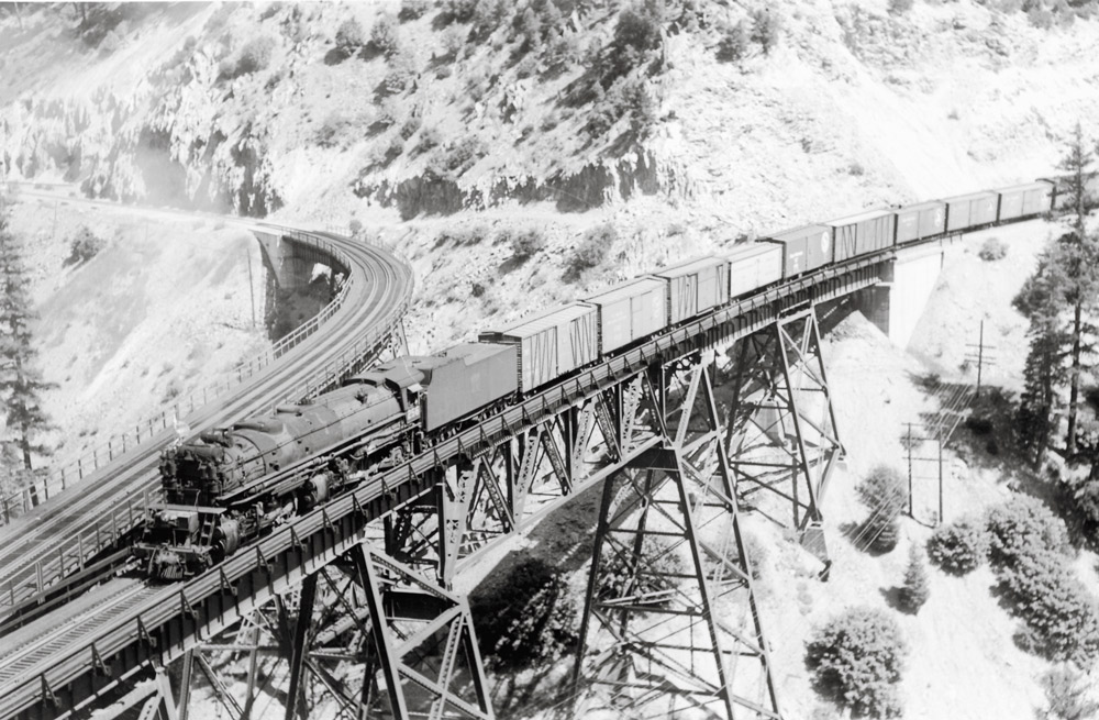 Black-and-white overhead view of steam locomotive with freight train on trestle in mountains