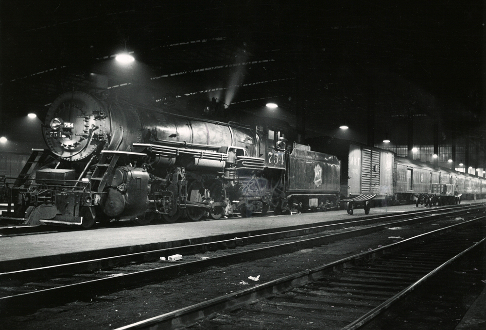 A steam locomotive and passenger train idle under platform lights next to a passenger platform at night.
