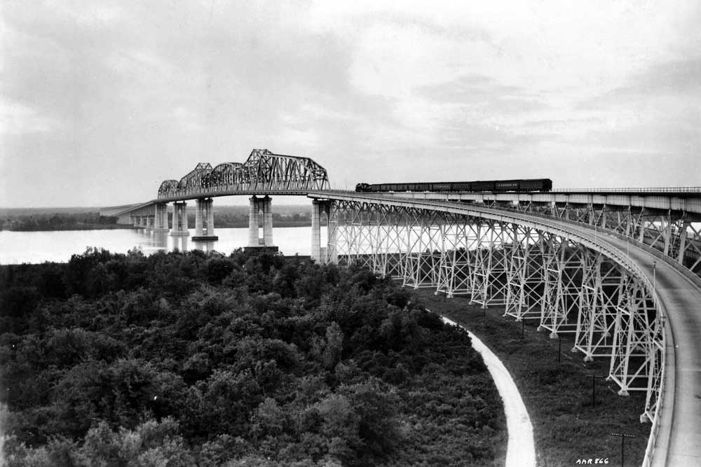 Black and white image of a passenger train crossing a trestle and bridge over a river and made small by the size of them all.
