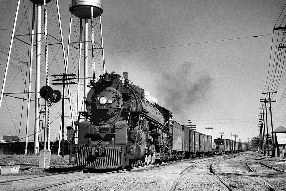 A large steam locomotive leads a freight train beside water towers under clear skies.