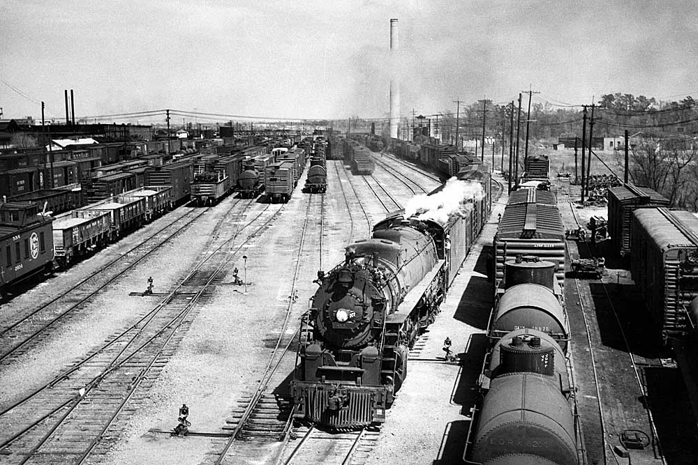 A steam locomotive hauling a freight train out of a rail yard.