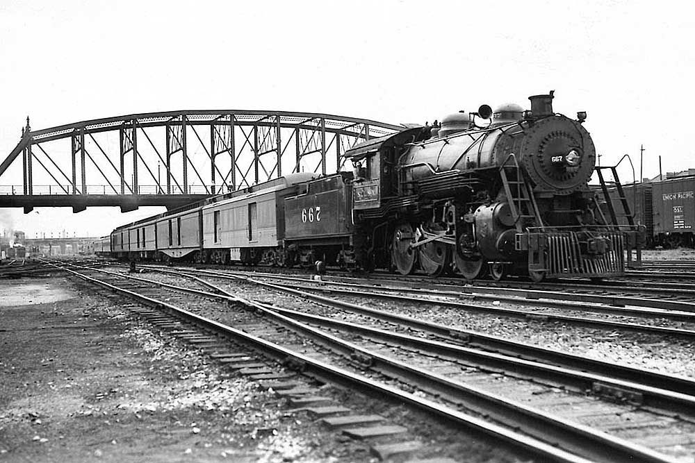 An older steam locomotive leads an express train underneath an arched girder bridge.