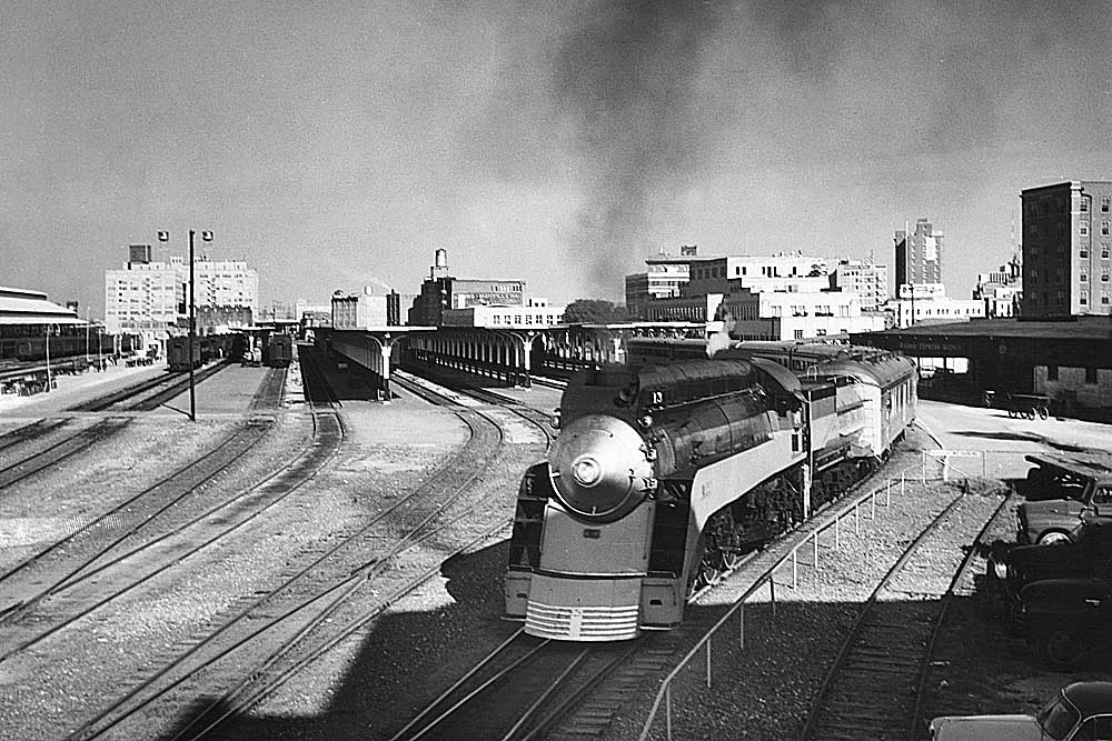 A streamlined steam locomotive leads a passenger train away from a passenger area toward the camera lens.