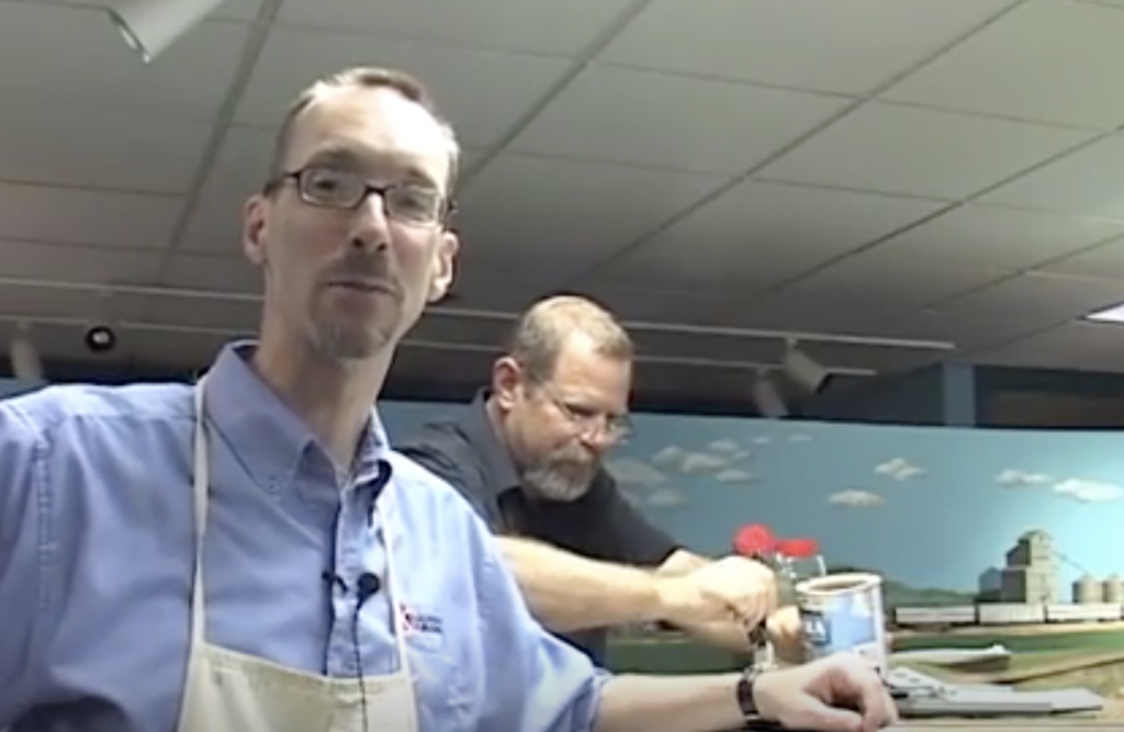 Two men appear next to a layout area to work on a model railroad.